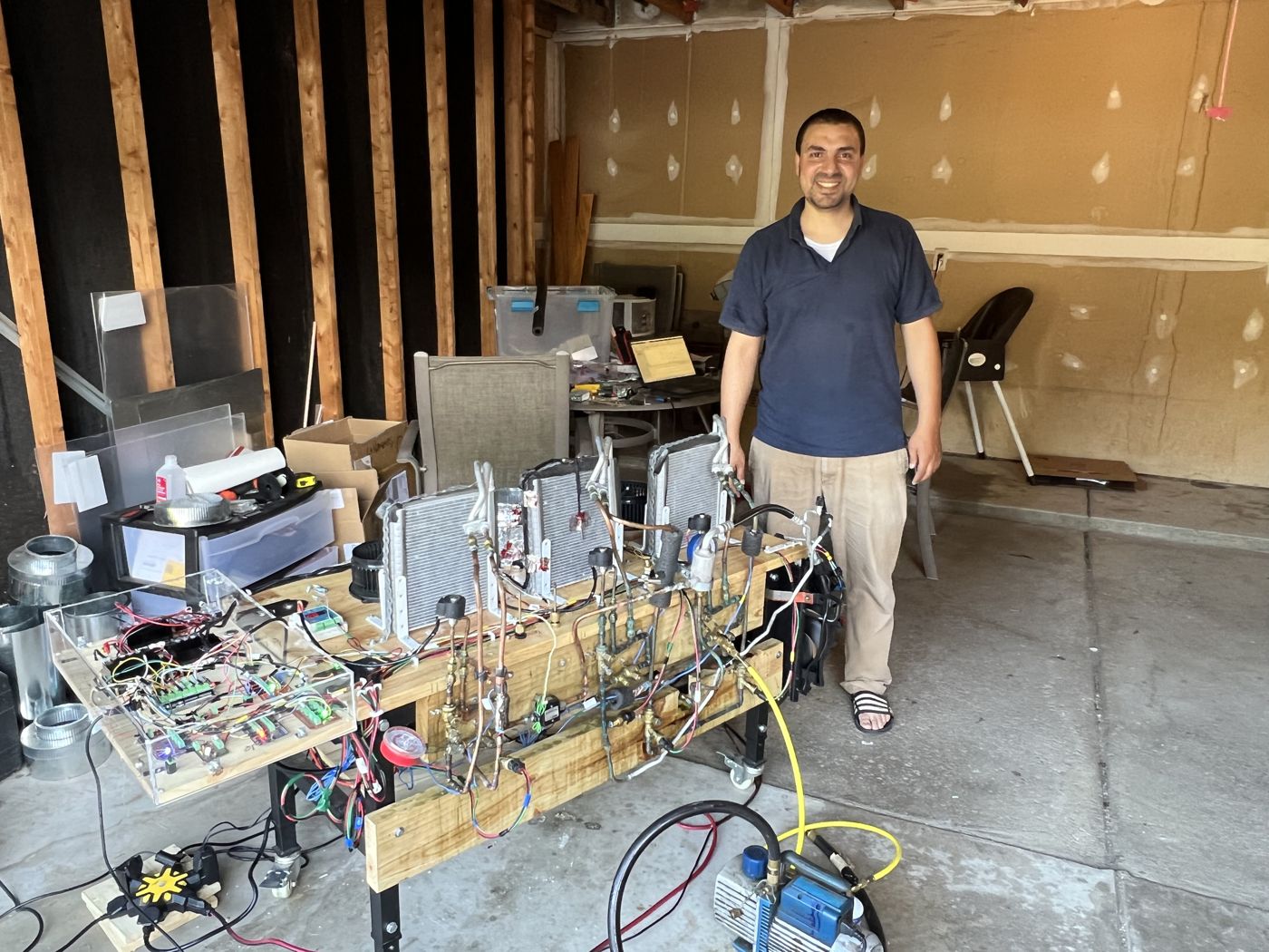 Ahmed standing next to the test bench: coils, blowers and copper lines on the workbench, with a clear box of controller electronics