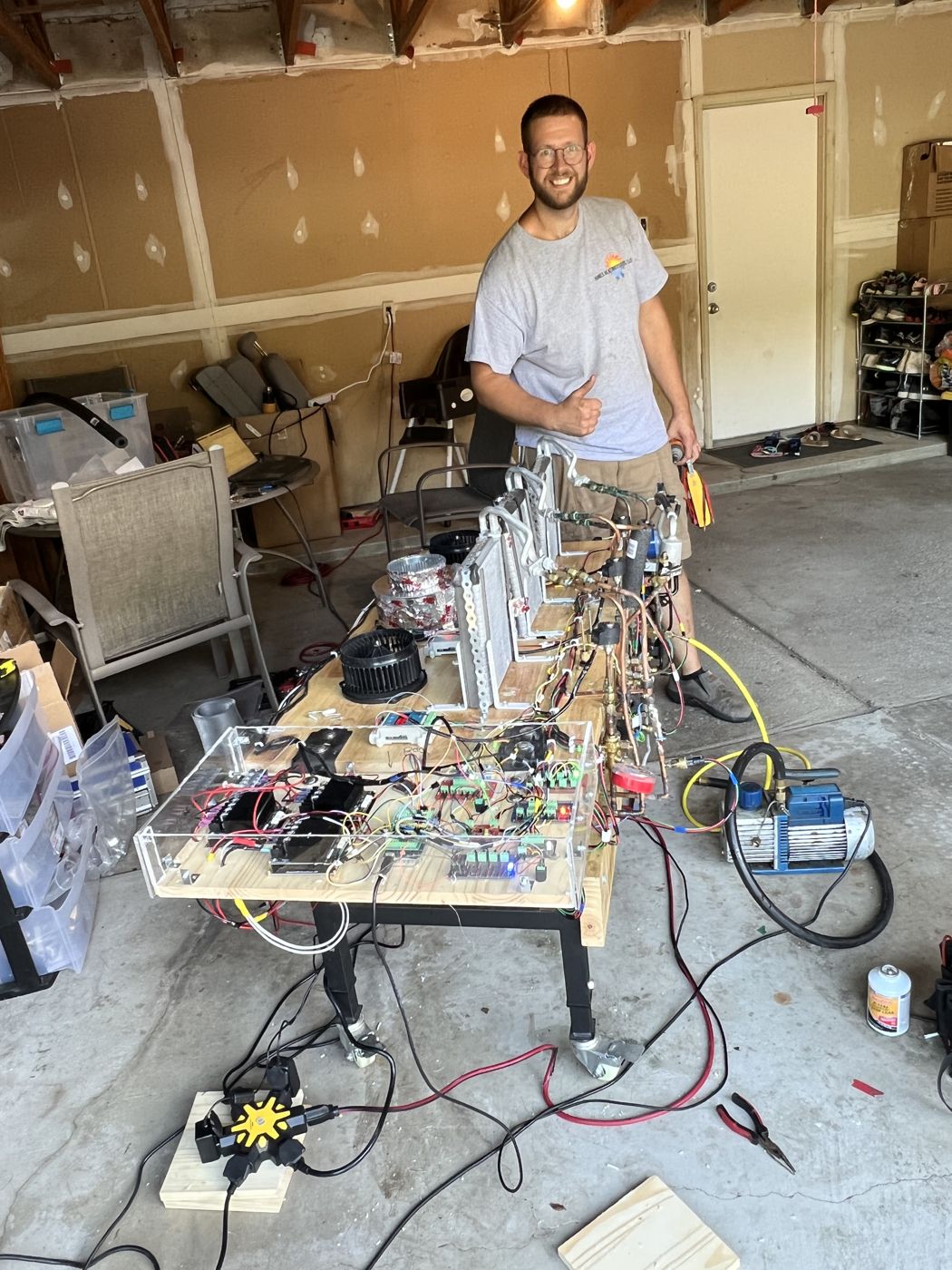 The AC technician at the bench giving a thumbs up, with a vacuum pump and refrigerant hoses on the garage floor