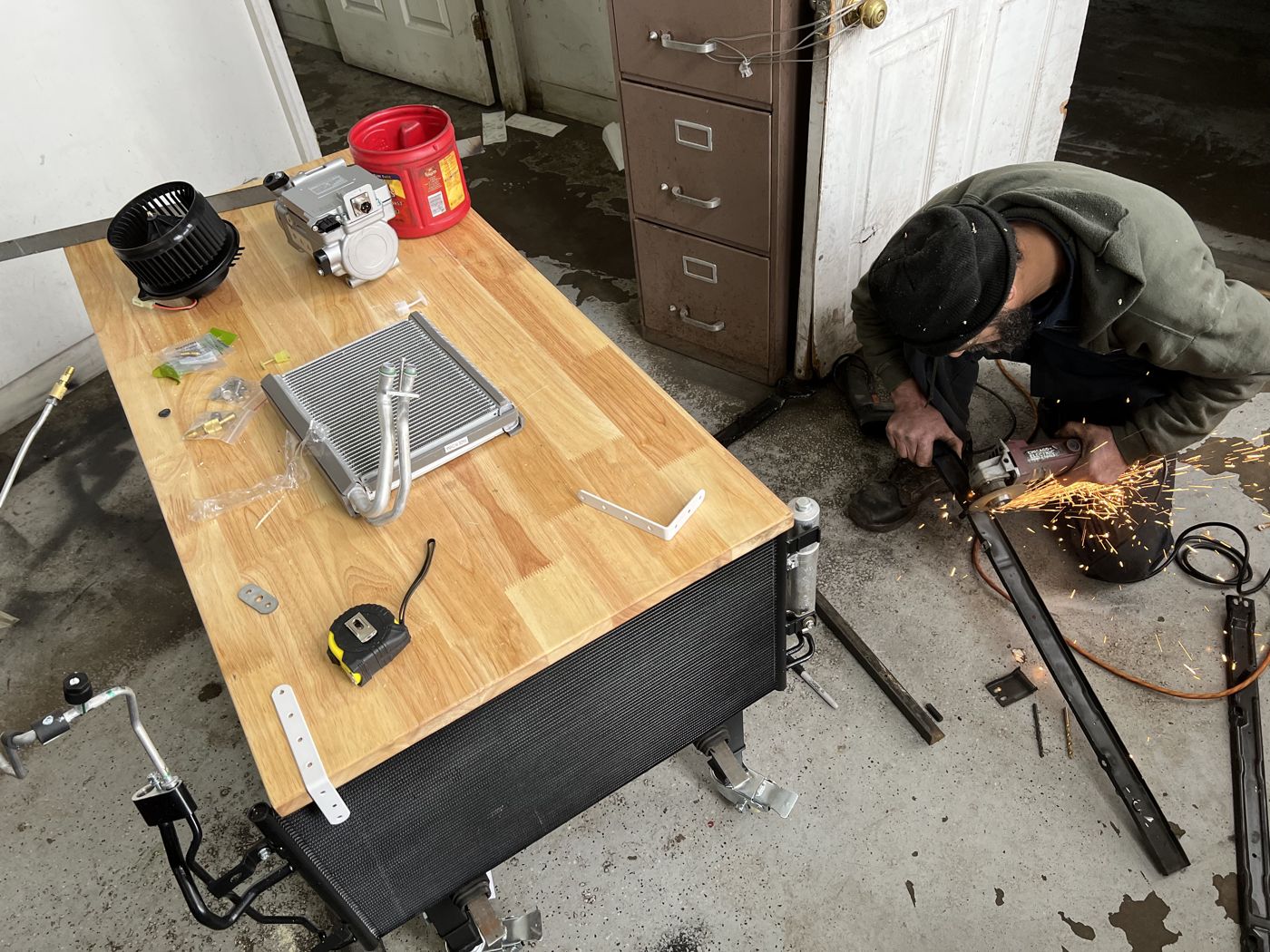 A mechanic cutting steel for the bench frame, beside a workbench holding a coil, a blower and a compressor